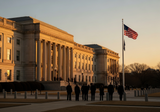 The exterior of the US State Department building at dusk with warm golden light, officials walking near the entrance, American flag visible