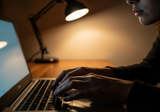 Close-up of hands typing on a laptop keyboard in a dimly lit room with screen glow and warm desk lamp